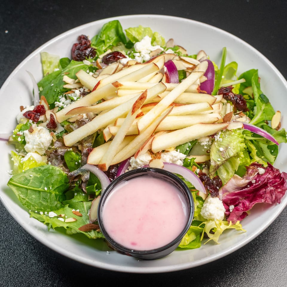 Fresh Italian salad with mixed greens, apple slices, goat cheese, cranberries, and a side of pink dressing.