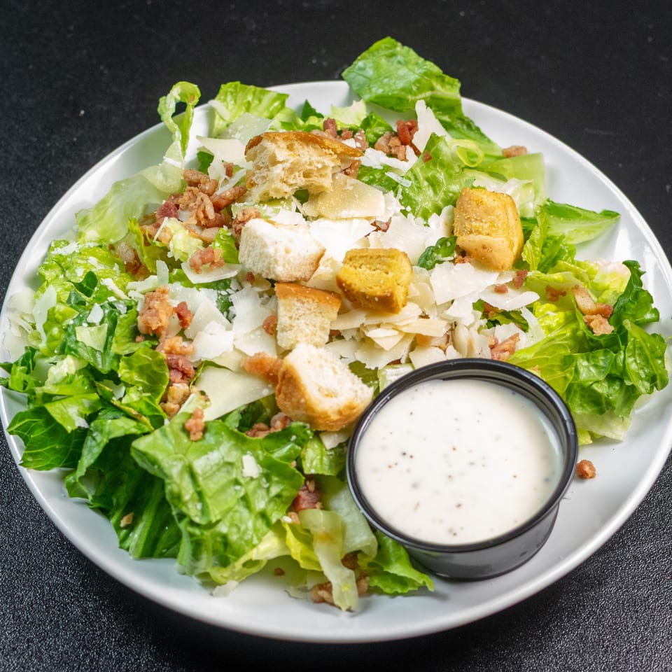 A Caesar salad with romaine lettuce, croutons, parmesan cheese, bacon bits, and a side of dressing.