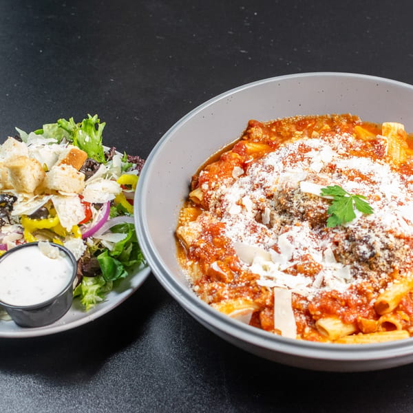 A bowl of rigatoni pasta with meat sauce and parmesan, next to a side salad with dressing.