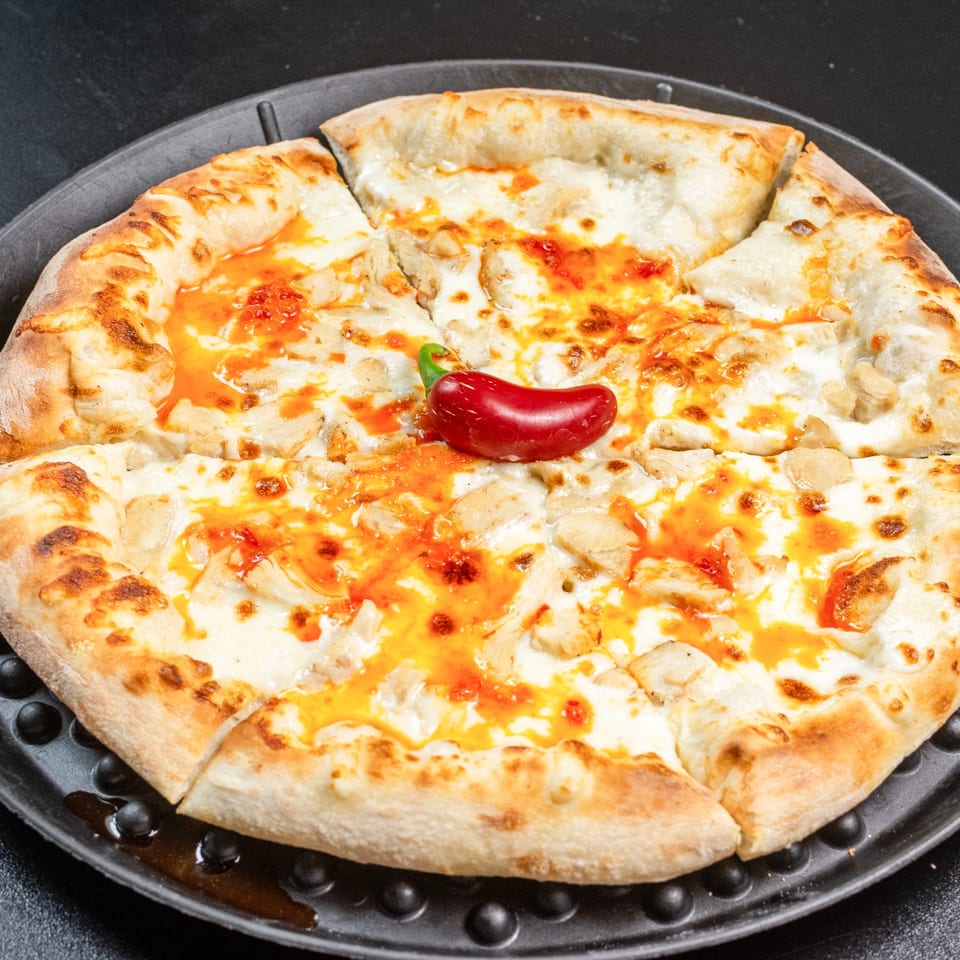 A whole chicken pizza with white sauce, orange drizzle, and a red chili pepper on top.